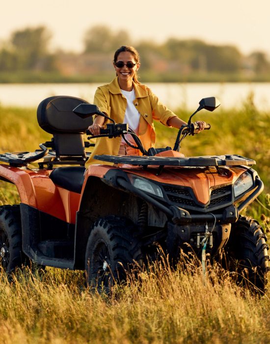 A cheerful brunette woman with sunglasses on driving a rented quad bike off-road, smiling for the camera.